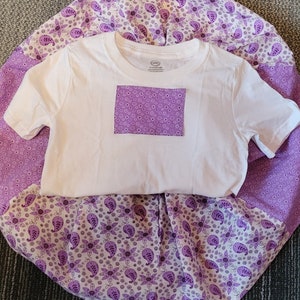 May include: A white t-shirt with a purple floral patch pocket on the front. The t-shirt is laying on top of a purple and white floral fabric.