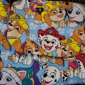 Op de afbeelding: Een lichtblauwe stoffen tas met een herhaald patroon van cartoonpuppy's. De puppy's zijn in tinten bruin, wit en geel, met hoeden en uniformen. De tas heeft een klepsluiting.