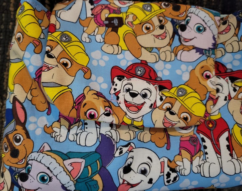 Op de afbeelding: Een lichtblauwe stoffen tas met een herhaald patroon van cartoonpuppy's. De puppy's zijn in tinten bruin, wit en geel, met hoeden en uniformen. De tas heeft een klepsluiting.