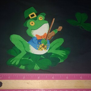 May include: A green frog wearing a green hat and a blue vest plays a violin. The frog is sitting on a green shamrock.