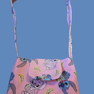 May include: A pink fabric shoulder bag featuring a repeating pattern of the character Stitch from Lilo & Stitch, along with yellow banana illustrations. The bag has a flap closure and a matching strap.