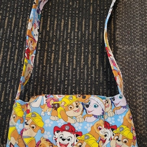 Op de afbeelding: Een rechthoekige schoudertas met een kleurrijke print van cartoonpuppy's op een lichtblauwe achtergrond. De tas heeft een bijpassende riem met hetzelfde puppy-patroon. De tas is gemaakt van stof en heeft een klepsluiting.