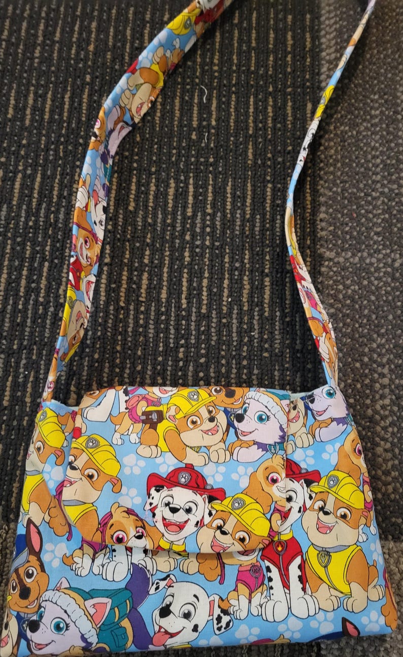 Op de afbeelding: Een rechthoekige schoudertas met een kleurrijke print van cartoonpuppy's op een lichtblauwe achtergrond. De tas heeft een bijpassende riem met hetzelfde puppy-patroon. De tas is gemaakt van stof en heeft een klepsluiting.