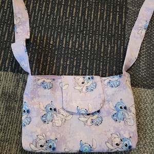 May include: A lavender crossbody bag featuring a repeating pattern of blue and white cartoon characters. The bag has a flap closure and matching lavender straps. The background has a speckled design.