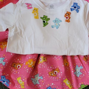 May include: A white t-shirt with a pink and white skirt. The t-shirt has a rainbow of Care Bears printed on the front. The skirt has a pink background with white stars and Care Bears printed on it.