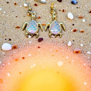 May include: A pair of gold-colored turtle earrings with iridescent shell-shaped centers. The earrings are displayed on a sandy surface with small shells and a blurred orange and yellow background.