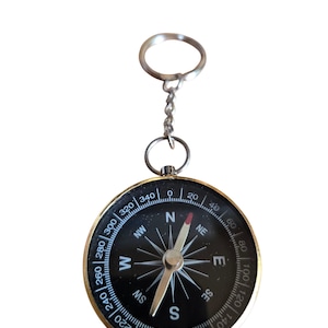 May include: A black compass with a gold-colored bezel and a silver keychain. The compass face features directional markings and a needle pointing north. The compass is a navigational tool.