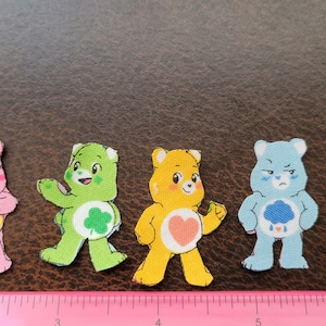 May include: Six Care Bears characters, each with a different color and design. The characters are arranged in a row from left to right. The characters are all smiling and have different expressions. The characters are all wearing different outfits. The characters are all made of fabric and have a soft, plush texture.