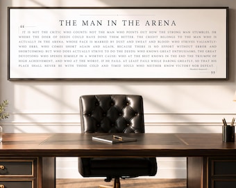 May include: A rectangular wall art piece featuring the text The Man in the Arena by Theodore Roosevelt in black serif font on a white background, framed in a thin dark border, displayed above a dark wood desk and a black tufted office chair.