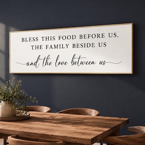 May include: A rectangular wall sign with a white background and a thin gold frame. The black text reads: BLESS THIS FOOD BEFORE US, THE FAMILY BESIDE US, and the love between us, with the final line in a decorative cursive script font.