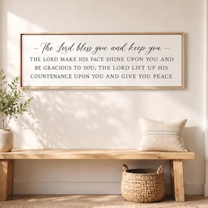 May include: A wooden bench with a woven basket, a decorative pillow, and a framed sign. The sign has the text "The Lord bless you and keep you" in black script. A vase with greenery is on the left.