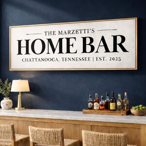 May include: A rectangular sign with the text "THE MARZETTI'S HOME BAR CHATTANOOGA, TENNESSEE | EST. 2025" in black lettering. The sign is framed in gold and hangs above a bar with woven bar stools. A lamp and bottles are also visible.