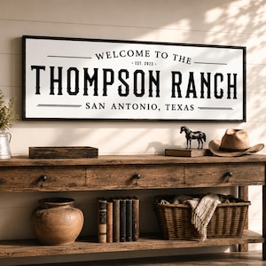 May include: A rectangular wall sign with a black frame and white background featuring the black text "WELCOME TO THE THOMPSON RANCH EST. 2023 SAN ANTONIO, TEXAS" in a bold, serif font, centered above a wooden console table with decor.