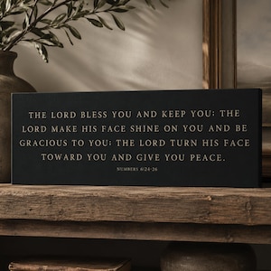 May include: A rectangular black sign featuring gold-colored text that reads: THE LORD BLESS YOU AND KEEP YOU; THE LORD MAKE HIS FACE SHINE ON YOU AND BE GRACIOUS TO YOU; THE LORD TURN HIS FACE TOWARD YOU AND GIVE YOU PEACE. NUMBERS 6:24-26.