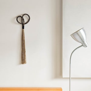 May include: A decorative wall hanging featuring two intertwined black and beige rope circles, connected to a long, beige tassel. The wall hanging is mounted on a white wall. A silver-colored lamp with a flexible neck is in the background.
