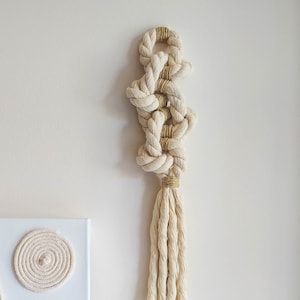 May include: Cream-colored macrame wall hanging with a knotted design and a tassel. The macrame is accented with natural-colored twine and hangs against a white wall. A canvas art piece with a spiral and curved design is visible on the left.
