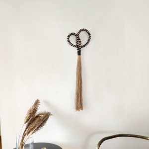 May include: A woven wall hanging with a heart-shaped top and a long, fringed bottom. The top is black and tan rope, and the fringe is a natural tan color. The wall hanging is displayed on a white wall.