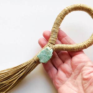 Könnte beinhalten: Ein handgefertigter Makramee-Ring aus natürlichem Jute-Seil mit einem hellgrünen Stein-Akzent. Der Ring ist unten mit einem Quasten gebunden.