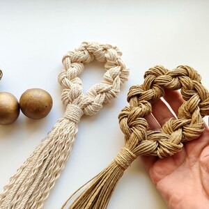 Puede incluir: Dos tapices de macramé para colgar en la pared, uno en beige y otro en marrón, con flecos de borlas. El tapiz beige tiene forma de círculo y el tapiz marrón tiene forma de corazón.