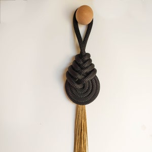 Puede incluir: Un tapiz de macramé de cuerda negra con una cuenta de madera en la parte superior y una borla larga y marrón.