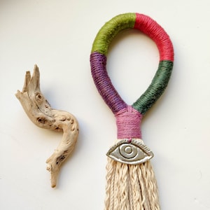Op de afbeelding: Een kleurrijke macramé wandhanger met een groene, roze en rode garenkwast. De kwast heeft een zilveren oogbedel en een witte katoenen franje.