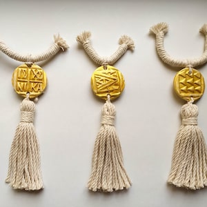 May include: Three gold tassel ornaments with geometric designs. Each ornament has a white tassel and a gold circular pendant with a different geometric pattern.