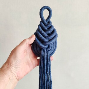May include: A blue macrame wall hanging with a tassel. The macrame is in the shape of a teardrop with a loop at the top.