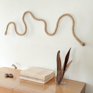 May include: A beige rope wall art piece in a wavy design is mounted on a white wall. Below, a stack of books, a glass vase with dried leaves, and a decorative metal sculpture sit on a wooden surface.