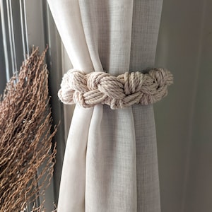 Puede incluir: Una cortina blanca y transparente con un amarre de cuerda beige grueso. La cuerda está trenzada y anudada alrededor de la cortina.