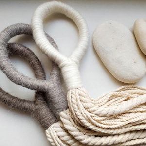 Può includere: Tre arazzi macramé in tonalità beige naturale e marrone. Gli arazzi sono realizzati con corda ritorta e hanno una frangia in basso.