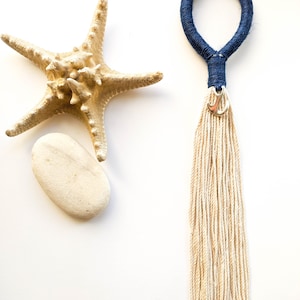 Op de afbeelding: Een sleutelhanger met een witte kwast, een blauwe lus van touw en een zilveren ankerbedel. De sleutelhanger ligt op een witte achtergrond met een zeester en een witte steen.