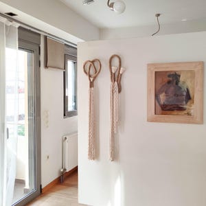 Può includere: Due arazzi da parete in macramè con un design annodato di colore marrone chiaro. Gli arazzi sono realizzati in corda di fibra naturale e sono appesi a una parete bianca. Un dipinto incorniciato con sfondo marrone è sulla parete a destra.