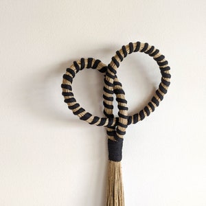 May include: A handcrafted wall hanging featuring two intertwined circular shapes, each wrapped with black and natural-colored rope. The design includes a central knot and a tassel at the bottom, creating a unique, textured decorative piece.