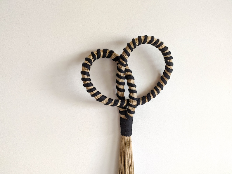 May include: A handcrafted wall hanging featuring two intertwined circular shapes, each wrapped with black and natural-colored rope. The design includes a central knot and a tassel at the bottom, creating a unique, textured decorative piece.