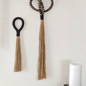 May include: Two woven wall hangings. The larger one has a figure-eight design with black and tan rope. The smaller one is a simple loop. Both have black wrapped handles and long, natural-colored tassels. Home decor.