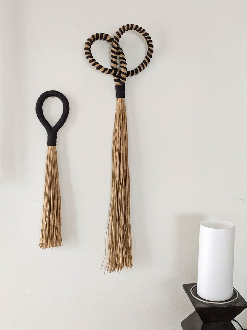 May include: Two woven wall hangings. The larger one has a figure-eight design with black and tan rope. The smaller one is a simple loop. Both have black wrapped handles and long, natural-colored tassels. Home decor.