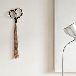 May include: A decorative wall hanging featuring a looped top with black and beige rope, connected to a long, natural-colored tassel. The design is minimalist and modern, suitable for home decor. The background is a plain white wall.