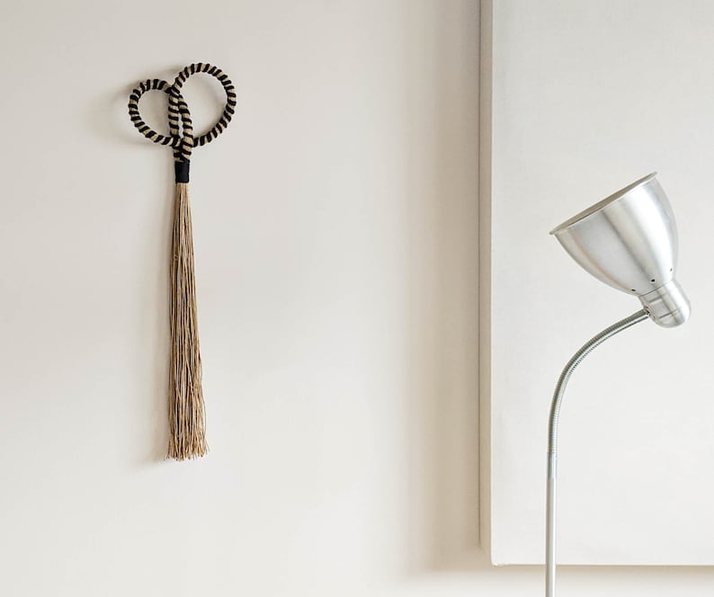 May include: A decorative wall hanging featuring a looped top with black and beige rope, connected to a long, natural-colored tassel. The design is minimalist and modern, suitable for home decor. The background is a plain white wall.