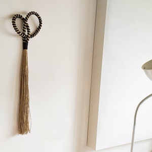May include: A decorative wall hanging featuring a heart-shaped design made of black and tan rope. The heart shape is connected to a long, flowing tassel made of natural-colored fibers. The piece is mounted on a white wall.