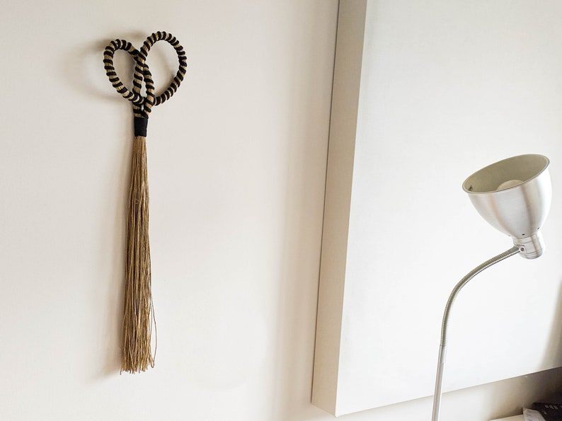 May include: A decorative wall hanging featuring a heart-shaped design made of black and tan rope. The heart shape is connected to a long, flowing tassel made of natural-colored fibers. The piece is mounted on a white wall.