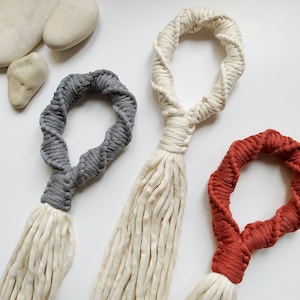 Könnte beinhalten: Drei Makramee-Pflanzenaufhänger in Grau, Off-White und Rostrot. Jeder Aufhänger hat eine gedrehte Schlaufe und eine lange, gefranste Quaste. Die Aufhänger werden auf einer weißen Oberfläche mit glatten Steinen präsentiert.
