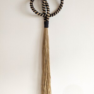 May include: A decorative wall hanging featuring two intertwined circular shapes with a black and tan rope design. The design includes a black band and a long, tan tassel, creating a unique, textured aesthetic.