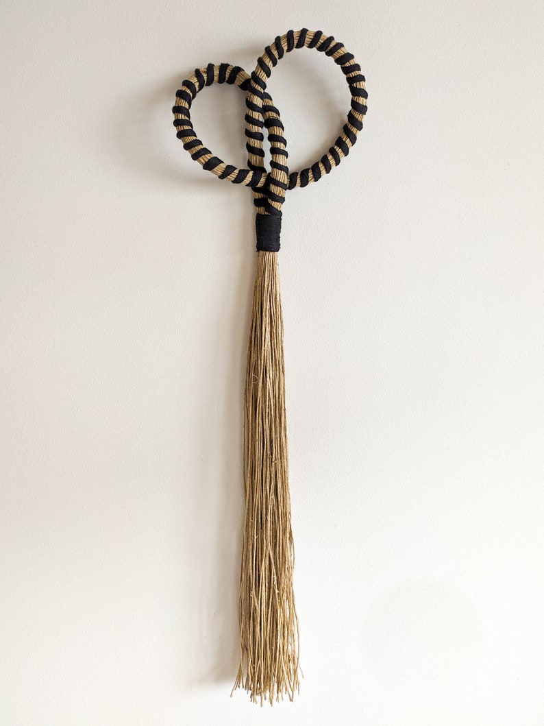 May include: A decorative wall hanging featuring two intertwined circular shapes with a black and tan rope design. The design includes a black band and a long, tan tassel, creating a unique, textured aesthetic.