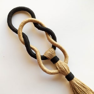 May include: A black and beige macrame wall hanging with a tassel. The macrame is made with two different types of cord, one black and one beige.