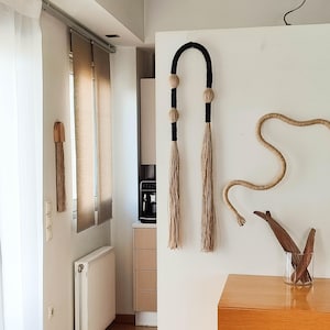 May include: Wall decor featuring two long, vertical pieces with black tops and beige tassels, and a curved rope design. A wooden piece with a tassel hangs on the left. The decor is against a white wall, with a window and wooden cabinet visible.