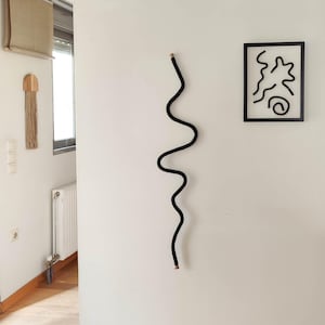 May include: A black, wavy wall sculpture with wooden ends hangs on a white wall. A framed abstract art piece and a wooden wall hanging with fringe are also visible. The sculpture adds a modern, minimalist touch to the space.