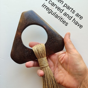 May include: A dark brown wooden triangle with a circular cutout and a long, natural fiber tassel hanging from the bottom. The wooden parts are hand carved and have irregularities.