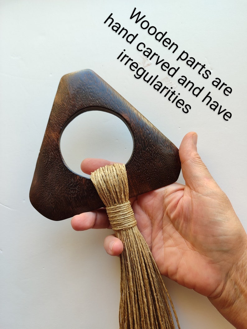 May include: A dark brown wooden triangle with a circular cutout and a long, natural fiber tassel hanging from the bottom. The wooden parts are hand carved and have irregularities.