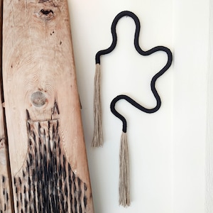 May include: Black macrame wall hanging with two long tassels. The wall hanging is shaped like a wavy line and is hanging on a white wall.