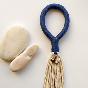 Puede incluir: Un círculo de cuerda azul marino con un borla y un dije de ancla dorada. El círculo de cuerda está colgado de una superficie blanca con dos piedras blancas.